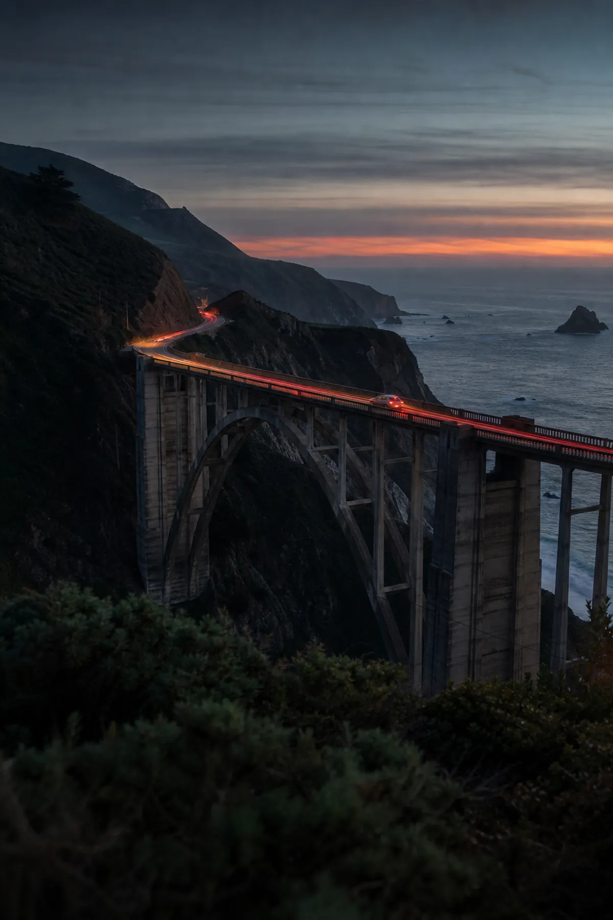 A car crossing Bixby Bridge at dusk, taillights tracing Highway 1